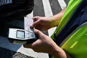 Policjant sprawdza prawo jazdy kierowcy, w dłoni trzyma także urządzenie do pomiaru prędkości.