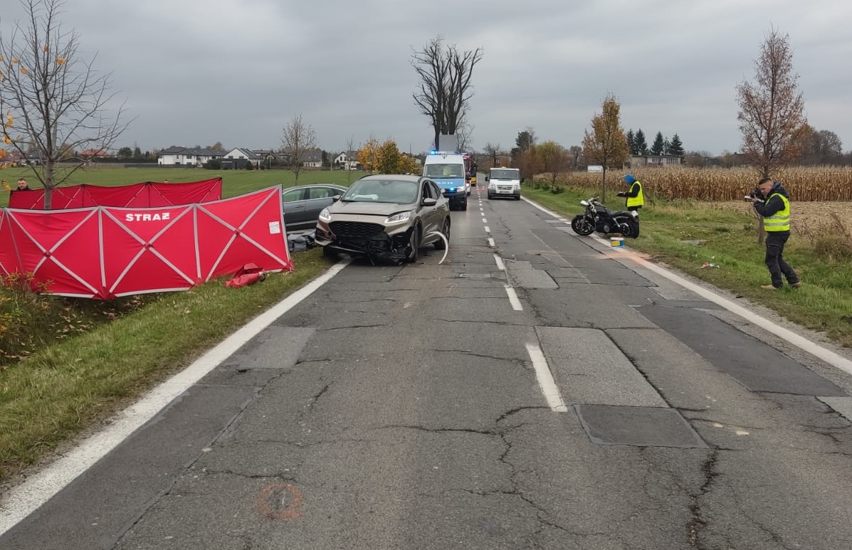 Miejsce wypadku drogowego. Po lewej widzimy uszkodzony samochód marki Ford Kuga, obok niego parawan strażacki. Po prawej widać motocykl uczestniczący w z zdarzeniu oraz policjantów dokumentujących - dokonujących oględzin.