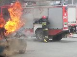 PO lewej płomienie wydobywające się z samochodu osobowego, w tle widać wóz strażacki i strażak sięgający po wąż...