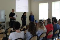Widzimy trójke policjantów. Od lewej policjant mężczyzna w mundurze, następnie policjantka w mundurze i po prawej policjantka po cywilnemu. Widzimy tez audytorium - słuchaczy.