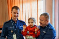 Policjanci umundurowani trzymają między sobą 5 letniego chłopca z odznaka policyjną.