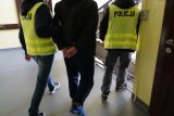 Dwaj policjanci w żółtych kamizelkach z napisem policja prowadzą skutego kajdankami z tyłu młodego mężczyznę.