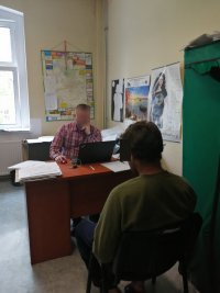 Plecami do nas podejrzany. Siedzi na krześle przed biurkiem. Za biurkiem przy komputerze mężczyzna (w domyśle śledczy) po cywilnemu.