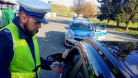 W tle radiowóz oznakowany. Na pierwszym planie policjant w białej czapce drogówki kontroluje kierowcę siedzącego za kierownica samochodu.