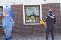 Policjant z tubą-megafonem i pies Sznupek w tle okna i dzieci.