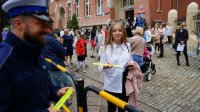 Po lewej usmiechniety policjant wręcza odblask dziewczynce. W tle tłum dzieci i rodziców przed szkołą.