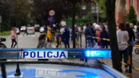 Na pierwszym planie radiowóz. W tle tłum ludzi przed szkołą, wśród nich policjant.