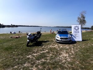 Plaża na której po lewej stoi policyjny motocykl, po prawej radiowóz policyjny i baner z napisem "komenda miejska policji w Gliwicach". W tle jezioro.