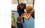 Policjant i nauczycielka pochylaja sie do dzieci i prezentuja im numer 112
