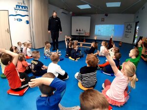 Dzieci w przedszkolu z podniesionymi raczkami - zgłaszaja sie do odpowiedzi. W tle policjant prowadzący zajęcia.