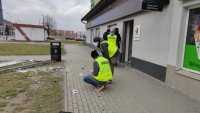 Policjanci prowadzący oględziny miejsca wysadzenia bankomatów. Dwóch techników kryminalistyki pochyla się nad czymś leżącym na chodniku. Trzech innych stoi przy drzwiach do budynku placówki bankowej.