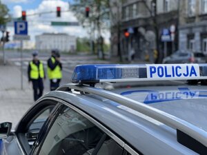 Policyjny radiowóz z widocznym "kogutem" - belką z napisem Policja, w tle dwoje policjantów, dalej przejście dla pieszych.