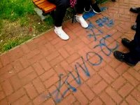 Widać buty siedzących na ławce wandali, napis na trotuarze i buty strażników miejskich.