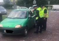 Policjant i Sokista rozmawiają z kierowcą, stoją przed szlabanem kolejowym.