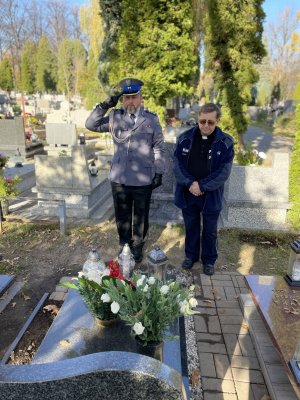 Policjant po lewej oddaje honory salutując. Po prawej stoi ksiądz. Obaj panowie stoją przy nagrobku obstawionym kwiatami i zniczami.