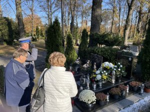 Oficer salutuje, stojac naprzeciwko nagrobku policjanta. Obok stoi matka pochowanego policjanta i ksiądz.