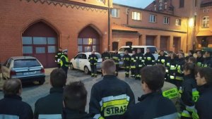 Policjant szkoli kilkudziesięciu strażaków na dziedzińcu komendy straży pożarnej.