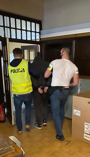 Dwaj policjanci po cywilnemu prowadzą korytarzem w komisariacie zatrzymanego meżczyznę.