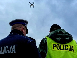 Dwaj policjanci zwróceni do nas plecami. W tle na niebie widać unoszącego się drona.