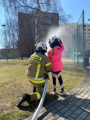 Strażak i dziecko trzymają wąż strażacki i kierują strumień wody w stronę boiska.