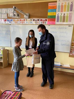 Policjant w klasie wręcza dziewczynce nagrodę za udział w konkursie plastycznym.