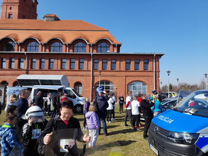 Piknik, wiele osób, szczególnie młodych, widać radiowóz policyjny, stoisko z materiałami profilaktycznymi, w tle budynek z cegły.