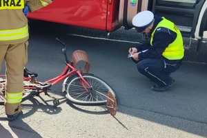 Po prawej kucający policjant, piszący notatkę, przed nim rower, który uczestniczył w wypadku (zgięte koło). Po lewej widzimy część stojącej postaci strażaka.