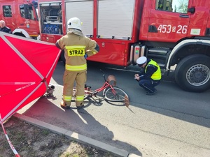 Od lewej widzimy parawan strażacki, obok stojący strażak, następnie leżący na jezdni rower, po prawej kucający i piszący oględziny policjant. W tle wóz strażacki.