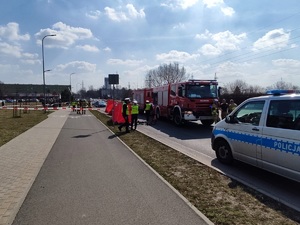 Po lewej chodnik, ścieżka rowerowa, po prawej na jezdni parawan, wóz strażacki  i radiowóz. Widać krzątających się policjantów i strażaków.