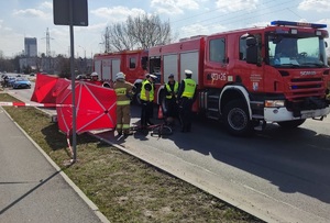 Po lewej fragment ścieżki rowerowej, po prawej na jezdni parawan, wóz strażacki. Widać krzątających się policjantów i strażaków.