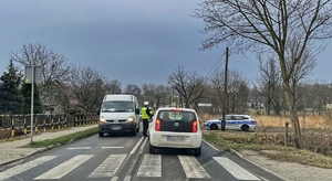 Widzimy jezdjnię z przejściem dla pieszych a za nim dwa samochody i dwoje policjantów dokonujących kontroli trzeźwości. Po prawej widać radiowóz.