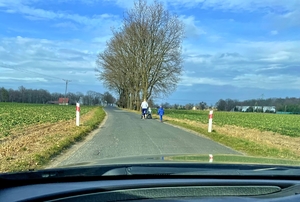 Wąska droga asfaltowa biegnąca przez pola rolne, po prawej stronie widać kobietę pchającą wózek, obok niej dziecko na rowerku biegowym i drugie starsze ok. 5 letnie - pieszo.