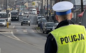 Policjant drogówki stoi po prawej i patrzy na przejścia na pieszych i ruch drogowy. W tle ulica - ruchliwa.