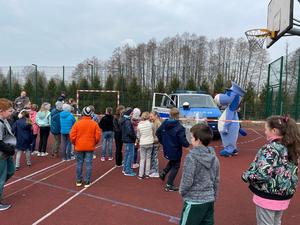 Dzieci szkolne na boisku przyszkolnym, widoczny policyjny radiowóz i policyjną maskotkę pies Sznupek (osoba przebrana za pluszowego psa).