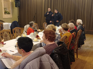 Sala bankietowa, widać stoły i siedzących przy nich seniorów, na drugim planie dwaj dzielnicowi.