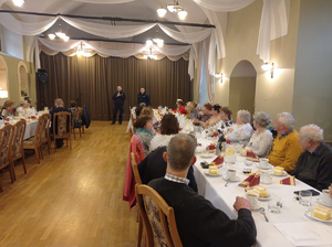 Sala bankietowa, widać stoły i siedzących przy nich seniorów, na drugim planie dwaj dzielnicowi.