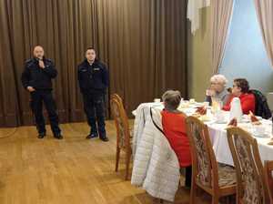 Sala bankietowa, widać stoły i siedzących przy nich seniorów, na drugim planie dwaj dzielnicowi.