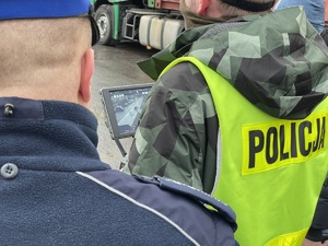 Dwaj policjanci, jeden z nich w kamizelce z napisem policja trzyma manipulator drona z ekranem - podglądem przejścia dla pieszych.