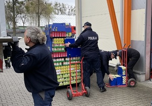 Na zdjęciu osoby rozładowujące paczki z towarami z samochodu ciężarowego. W centralnej części policjant przenosi paczki z artykułami spożywczymi.