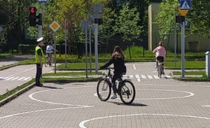 Miasteczko ruchu drogowego, po lewej stoi policjant i dogląda przebieg egzaminu praktycznego, dzieci jada rowerami.