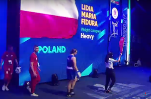 Trzy osoby w tym po środku Lidia fidura wchodzą na ring. W tle plansza z napisem POLAND Lidia Maria Fidura i flaga Polski.
