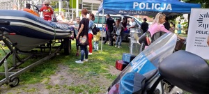 Policyjne stoisko podczas rodzinnego festynu, widać policyjna motorówkę, przód motocykla i policyjny furgon. Widać matki z dziećmi zwiedzające stoisko.