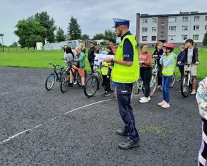 Policjant ruchu drogowego stoi na boisku, za nim dzieci na rowerach przygotowujące się do egzaminu na kartę rowerową.