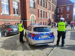 Policyjny radiowóz i dwóch policjantów po bokach przed budynkiem szkoły.