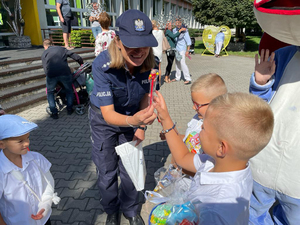 Policjantka wręcza pierwszoklasistom upominek. Rzecz dzieje się przed szkołą.