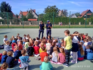 Dwaj policjanci prowadzą pogadankę dla grupy uczniów. Rzecz dzieje się na boisku szkolnym. Policjanci stoją, dzieci siedzą zasłuchane.