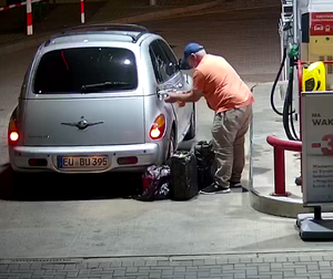 Tęgi mężczyzna tankuje kanistry na stacji benzynowej. Obok stoi zaparkowany samochód na niemieckich numerach rejestracyjnych.