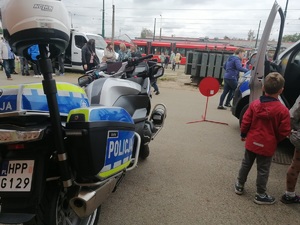 Po lewej stronie jest ustawiony motocykl policyjny stojący tyłem, po prawej stronie widzimy uczestnika festynu, w centralnej części zdjęcia w oddali widać zajezdnię tramwajową.