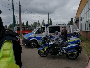 Zdjęcie przedstawia flotę gliwickiej Policji, stojący lewym bokiem radiowóz typu Van oraz przed nim stojący również bokiem motocykl, na którym siedzi dziecko. W oddali widać przejeżdżający tramwaj.