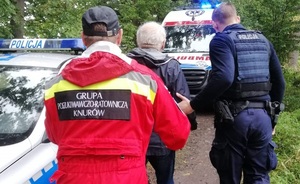 Na zdjęciu widzimy starszego mężczyznę prowadzonego przez ratownika z grupy poszukiwawczej i policjanta. W tle widać radiowóz i karetkę pogotowia.